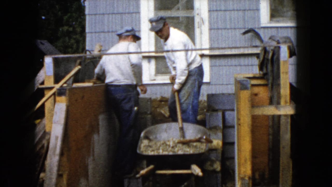 Two men shovel gravel near a wheelbarrow beside a shingled house.