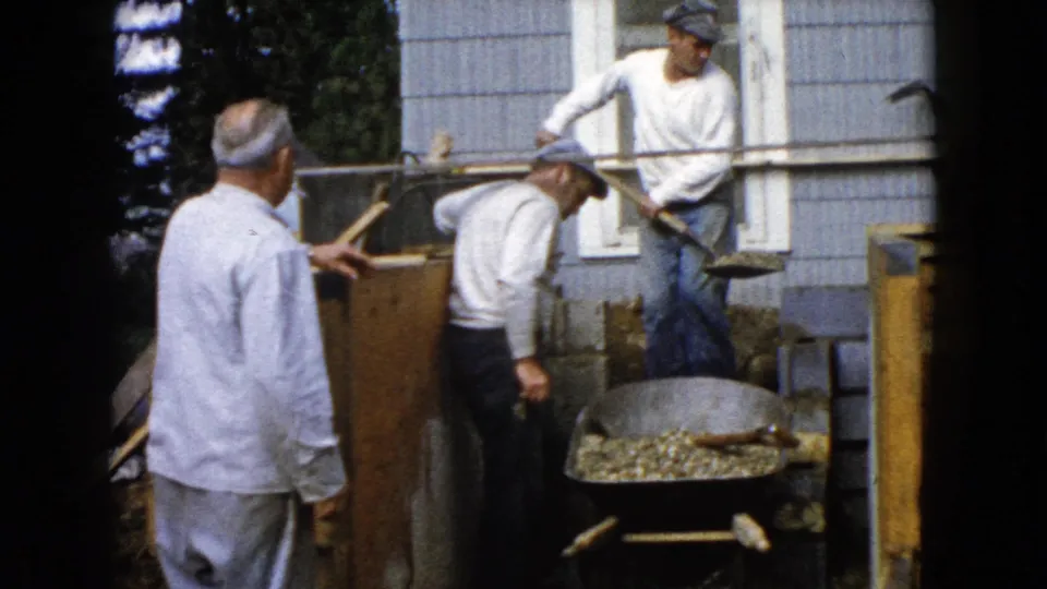A man in a light shirt watches as two others handle shovels near a wheelbarrow outside the Illinois house.