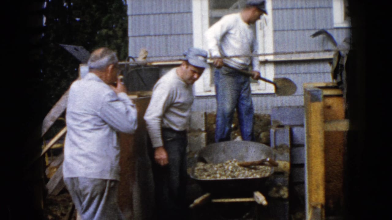 Several men work beside a house with shovels and a metal pan of concrete material.