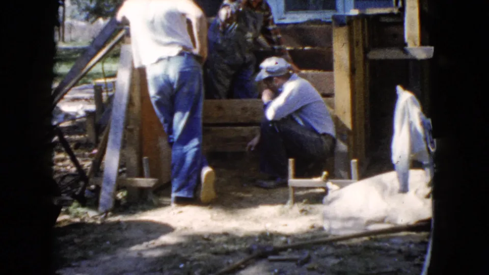 Adult men in caps and jeans gather near porch framing or steps at an outdoor residential work site in Wauconda, Illinois, in 1960.