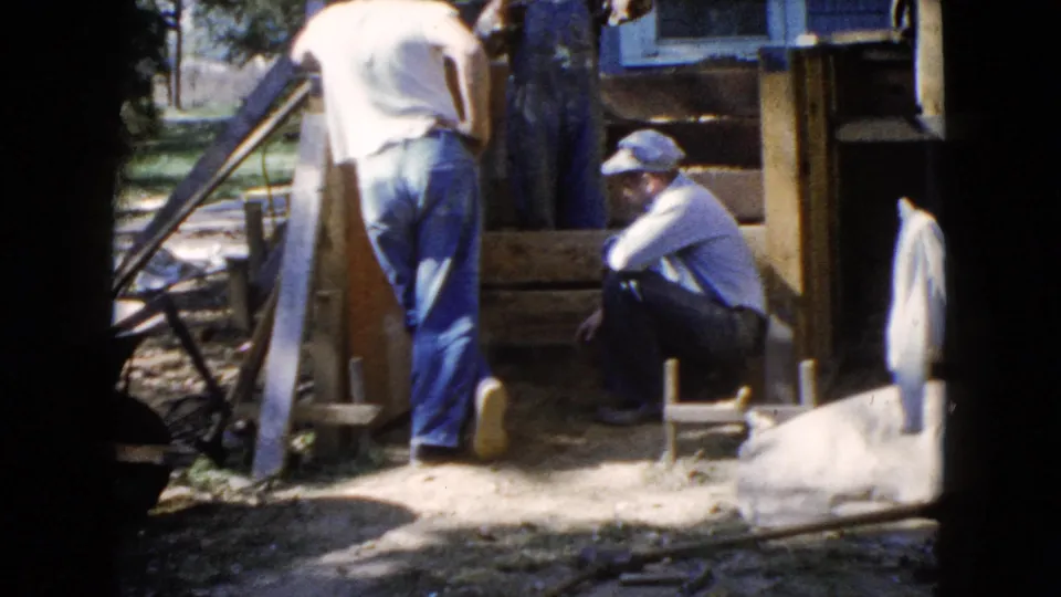 A 1960 home-movie frame shows men in casual workwear beside boards, steps, and house siding during a small residential construction task.