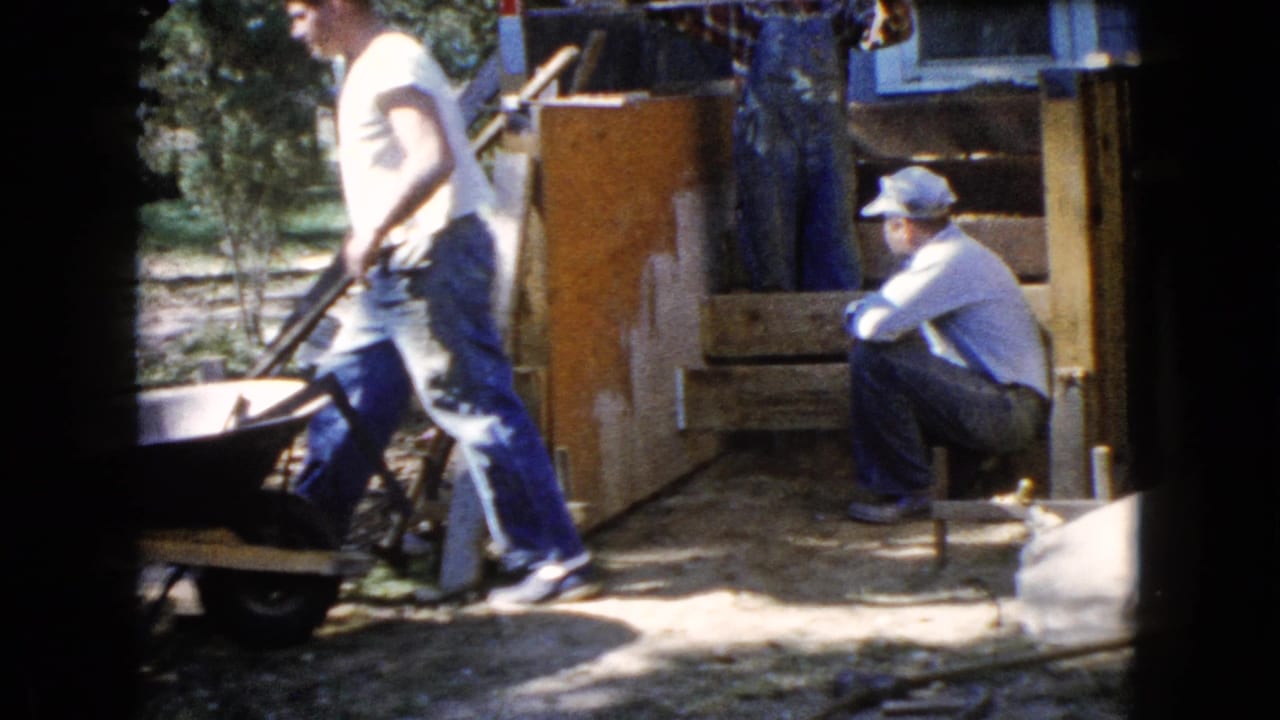 Men beside a wheelbarrow and wooden stair form in a sunny yard.