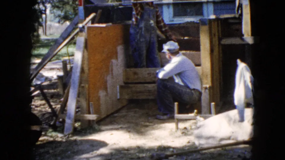 Home-movie frame of a seated man and a partially visible worker around wooden stair formwork in a sunny Illinois yard.