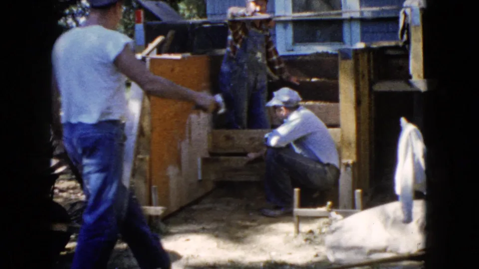 A man in a white T-shirt walks through the foreground while another sits near the stair form in 1960 Wauconda footage.