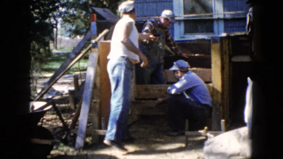 A man in a white tank top stands beside the deck frame as two other men work near the house exterior in Wauconda, Illinois.