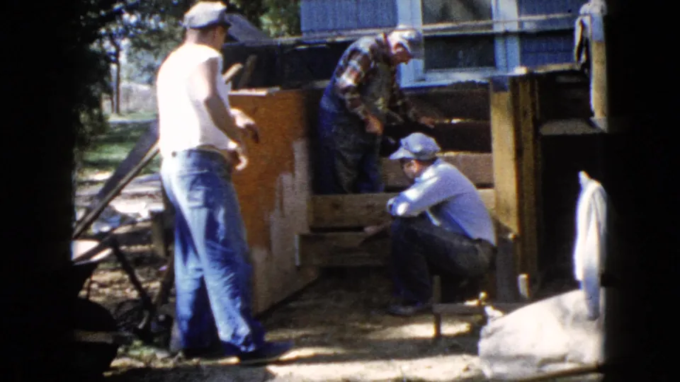The 8mm frame shows three adult men at a residential deck repair, with one crouched by the frame and another working from inside it.