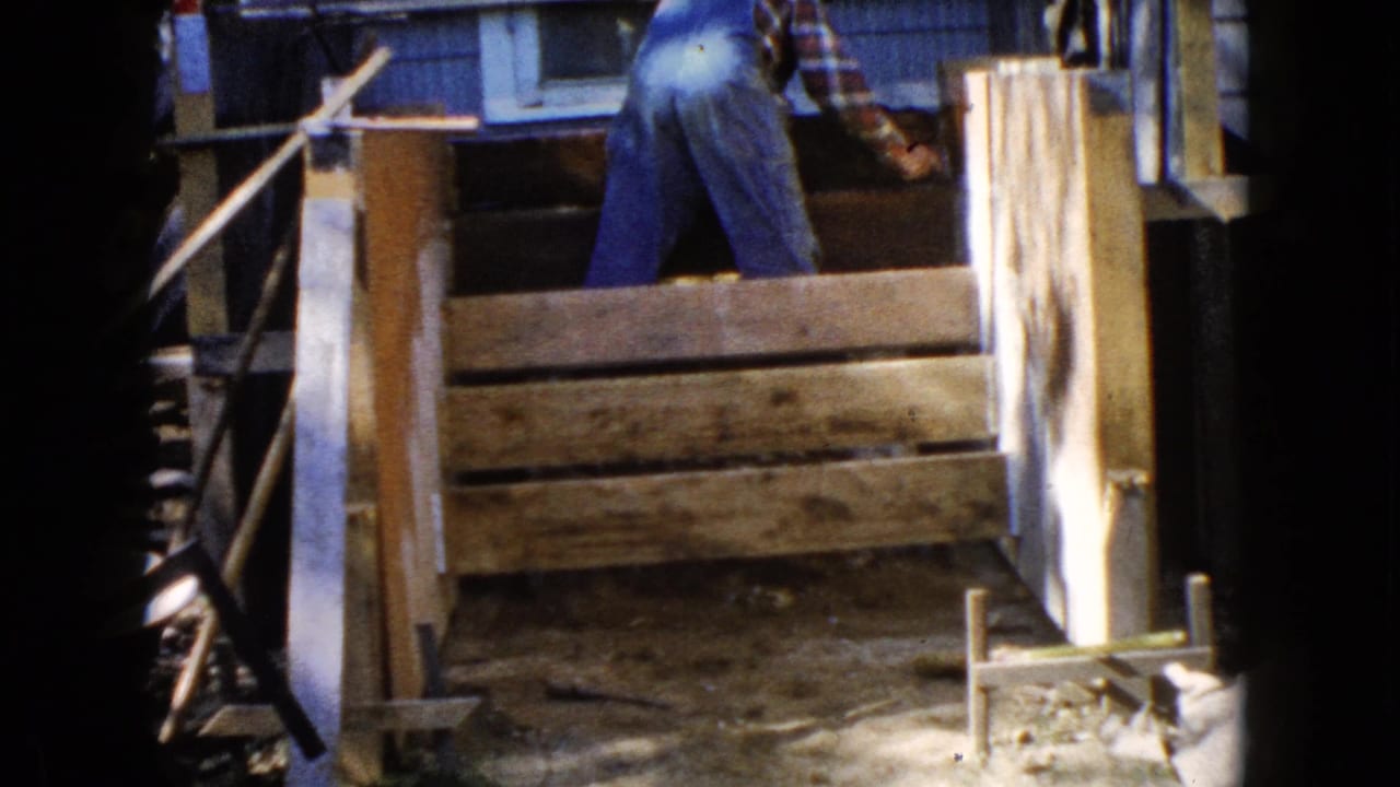 In Wauconda, Illinois, 1960 home-movie footage, a man in blue overalls works over a small wooden structure near house siding.