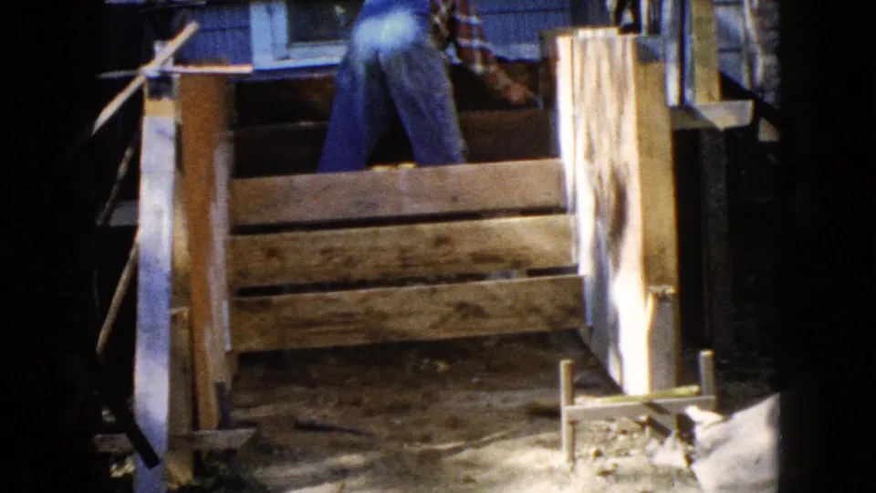 The 1960 Wauconda frame shows a man partially visible above a rough wooden frame, with house siding and a window behind him.