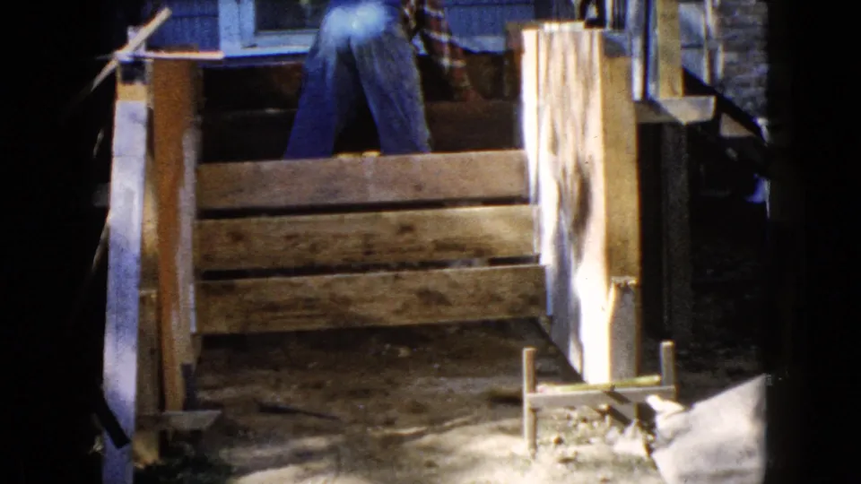 A 1960 home-movie frame from Wauconda shows rough boards and posts in front of a man working near the side of a house.