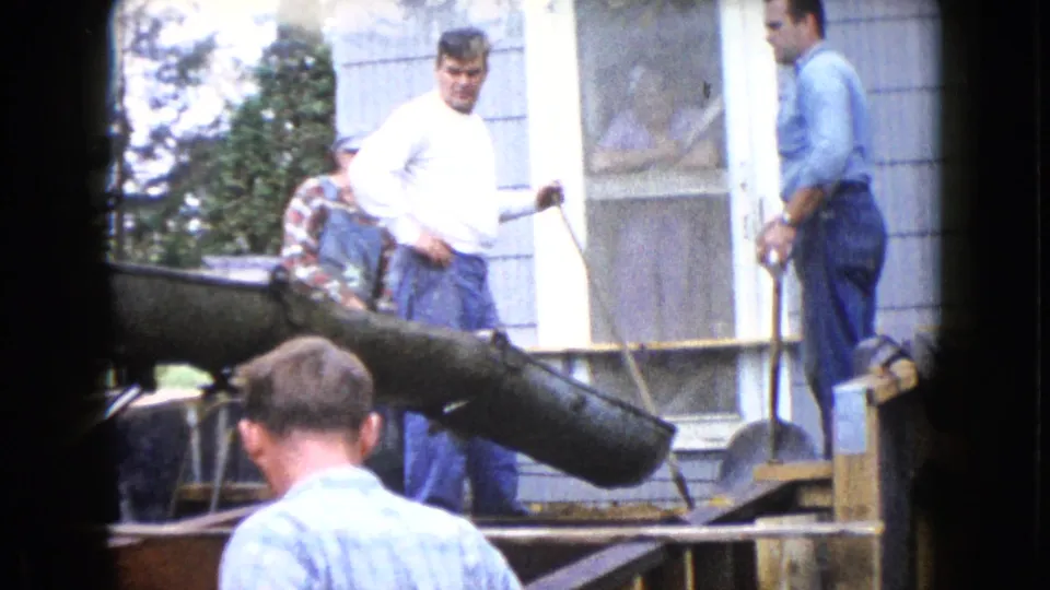 In Wauconda, Illinois, 1960, men in work clothes stand by a concrete chute beside a gray-sided house during construction.