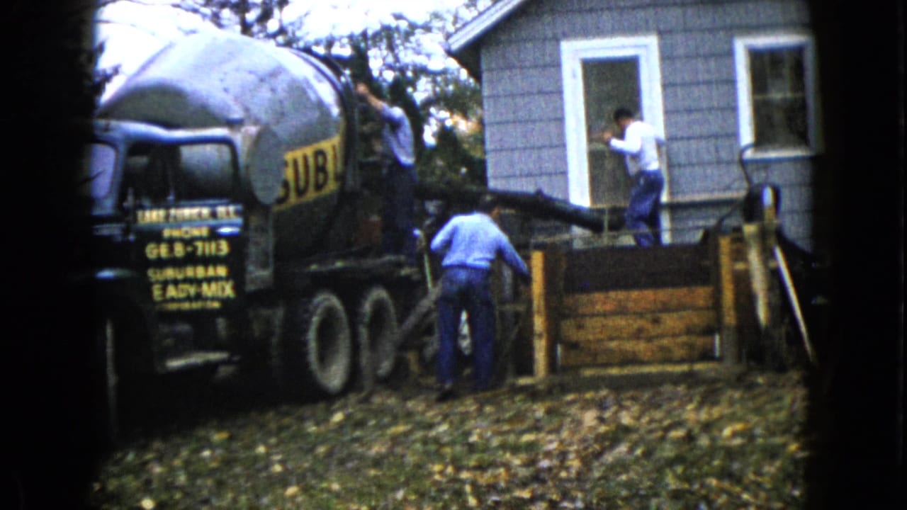 Cement mixer truck beside a gray house as men guide concrete toward a porch form.
