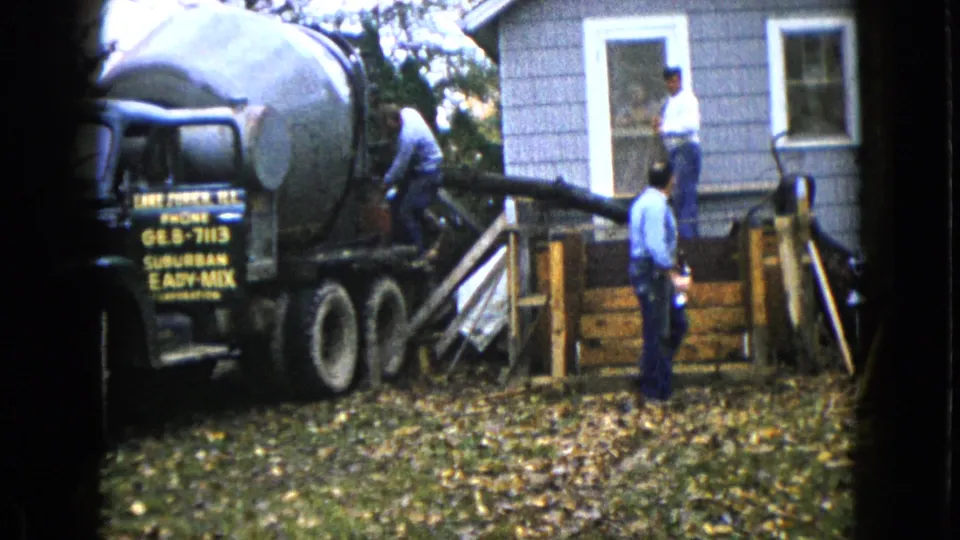 Workers in blue trousers stand near a wooden porch form while the cement truck chute reaches toward the house.