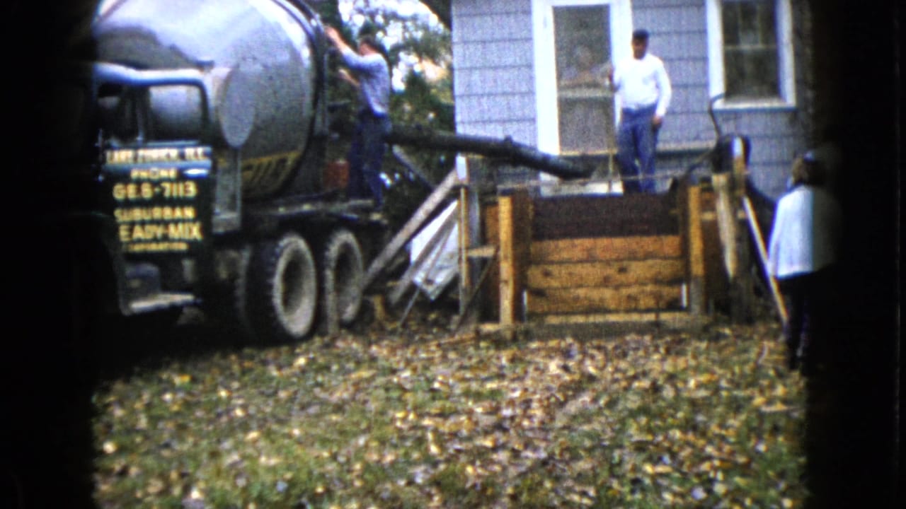 Cement mixer truck beside a house as workers prepare a concrete pour near wooden forms.