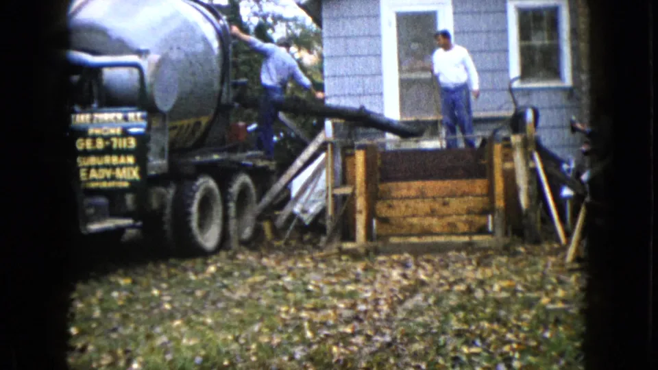 1960 Wauconda, Illinois frame with a concrete truck chute positioned toward wooden forms beside a gray-sided house.