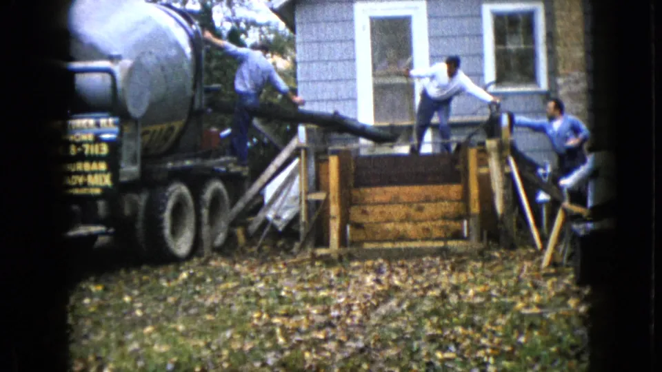 1960 Wauconda, Illinois home-movie frame showing three men coordinating a concrete pour from a mixer truck near porch steps.