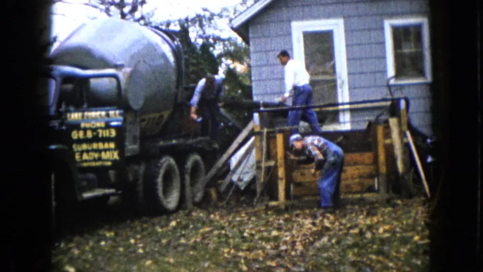 1960 Wauconda, Illinois frame showing workers repositioning concrete-pour equipment around a wooden form in a residential yard.