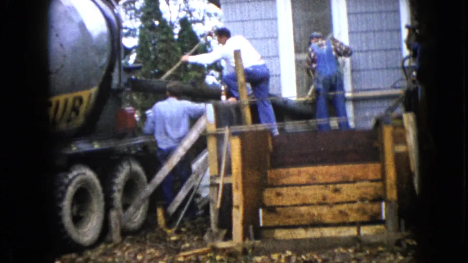 A worker leans toward the concrete chute while others stand beside wooden forms at a 1960 Wauconda residential construction site.