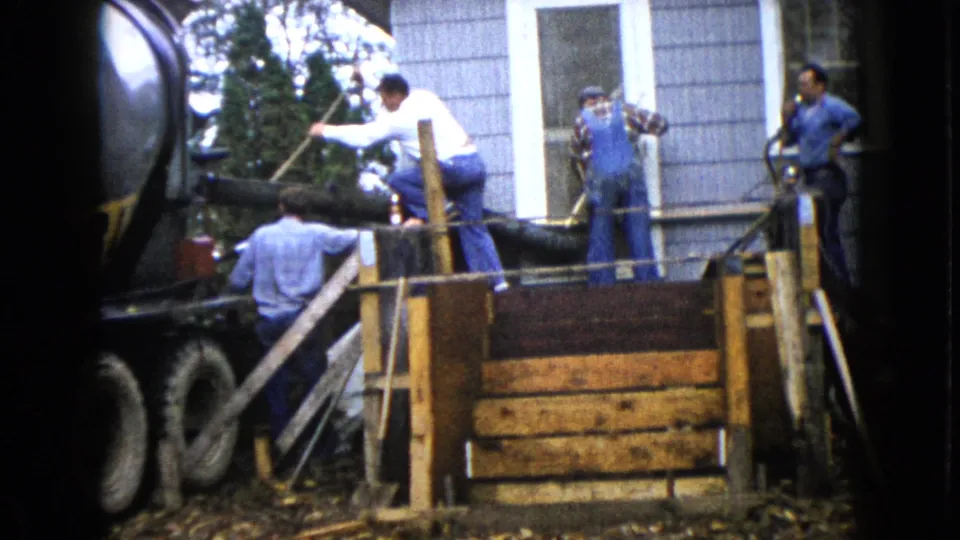 Men in work clothes continue a concrete pour beside a house, with a truck chute feeding wooden formwork in Wauconda, Illinois, 1960.