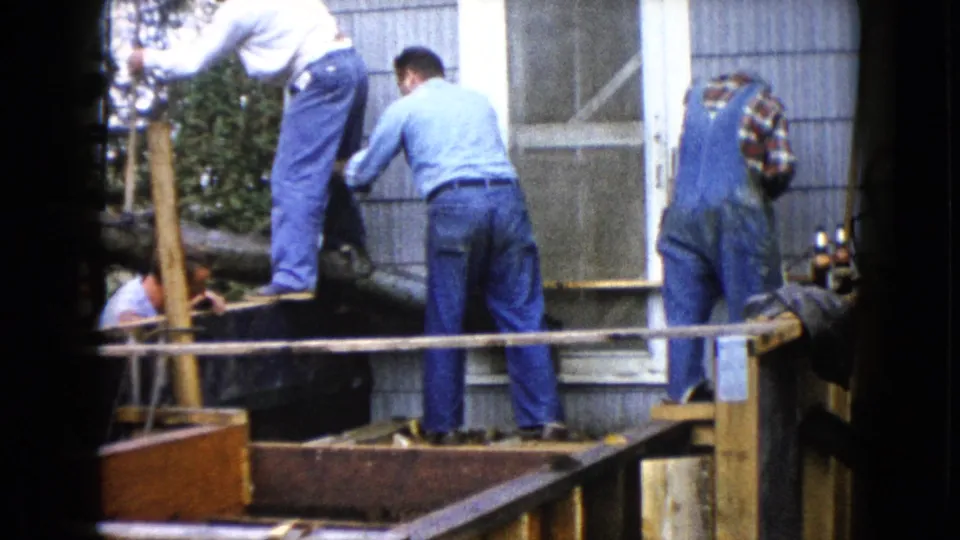 Closer 8mm view of men working beside a doorway as wet cement is leveled within the porch structure.