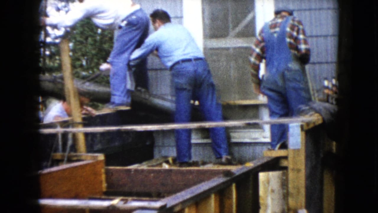 Men in jeans and overalls work around a wooden structure beside a gray-sided building.