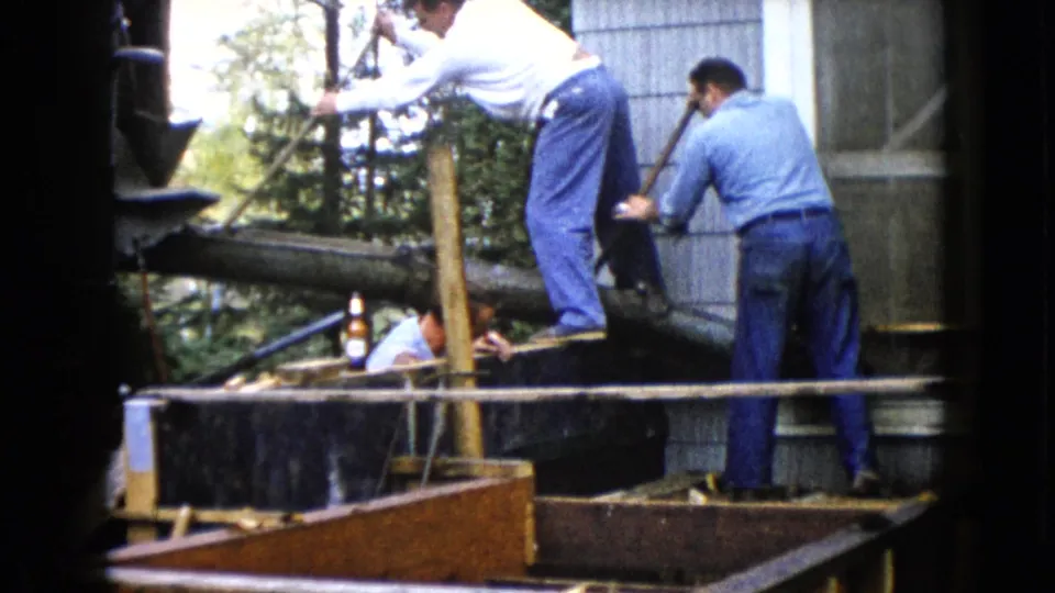 1960 Wauconda outdoor construction scene with men leaning over a beam beside a gray-sided building and trees.