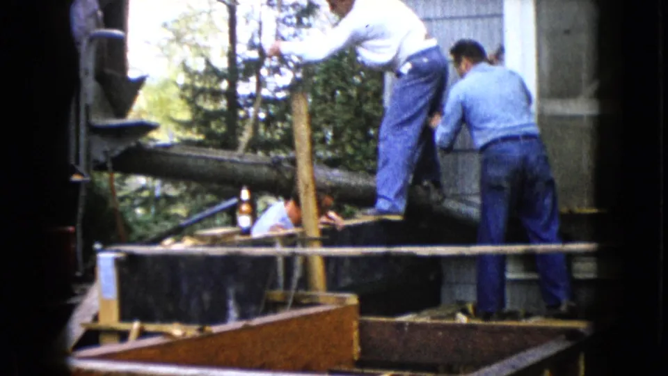 1960 Wauconda frame showing men in denim and work shirts handling wood on an outdoor building project.