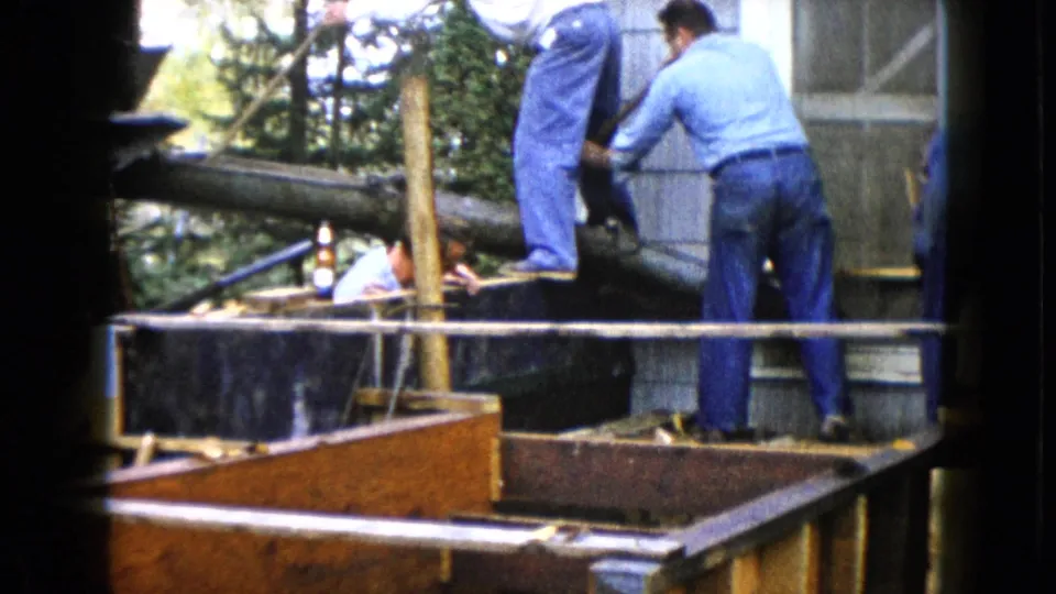 1960 Wauconda home-movie view of men working on a wooden outdoor structure beside a gray-sided building.