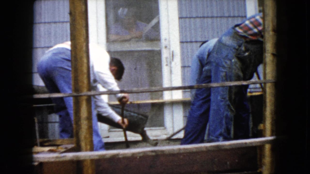 In 1960 Wauconda, Illinois, two men work by wood formwork outside a sided house, one leaning over a wheelbarrow of cement.