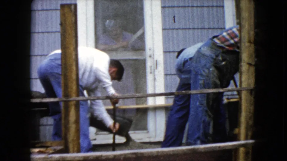 A 1960 Wauconda home-movie frame shows a man tipping a wheelbarrow of cement as another works near the wooden frame.
