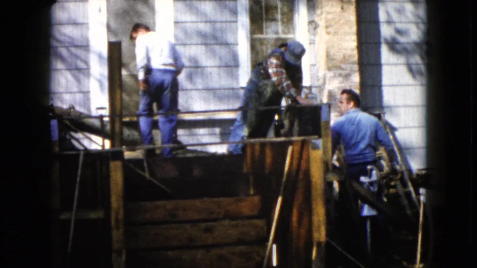 A 1960 home-movie frame shows men working on a wooden porch structure, with a ladder and house windows visible nearby.