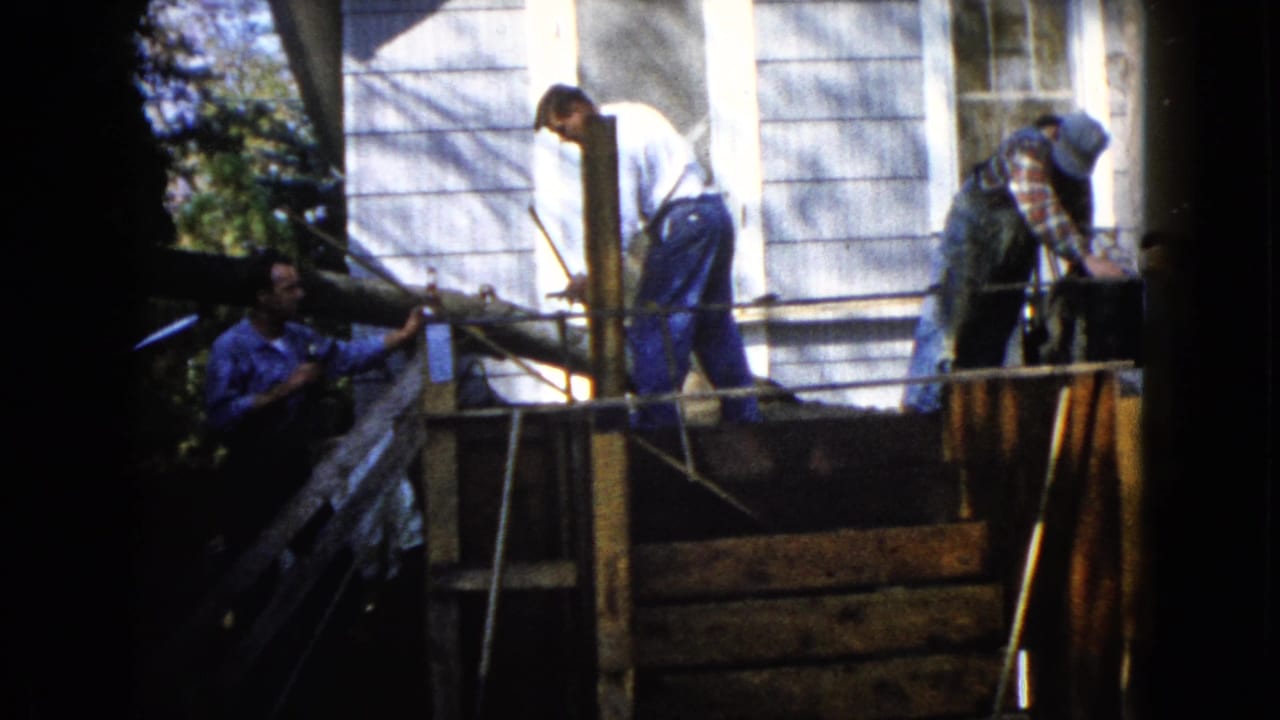Men work around a wooden platform beside a light-sided house, using ropes near a porch area.