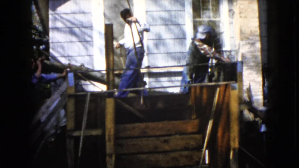 Two adult men stand on a raised platform by the house while another assists from below, with ropes visible in the Wauconda home-movie frame.
