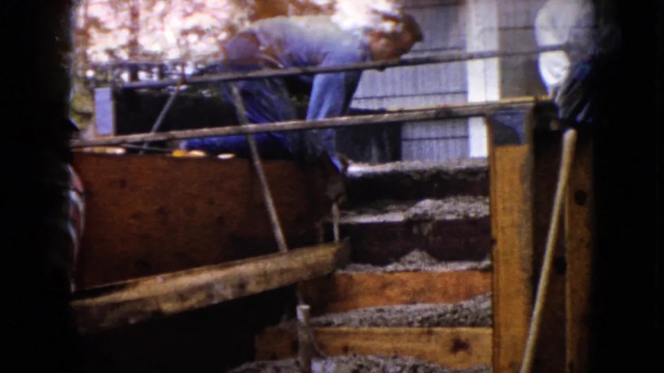 1960 Wauconda frame showing a man working on a wooden form or structure beside exterior steps of a house.
