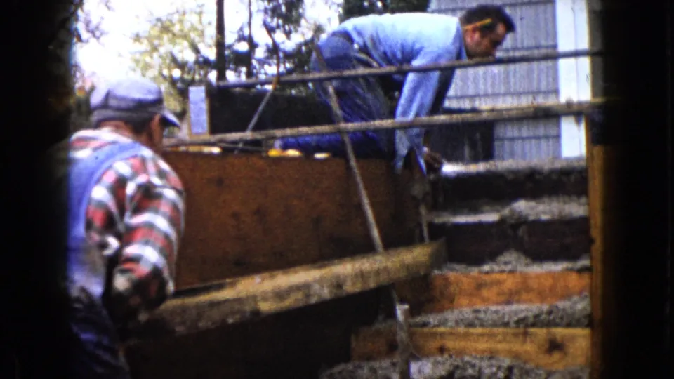 A worker in a blue shirt leans across wooden concrete forms as another man in a cap watches from beside the step area.