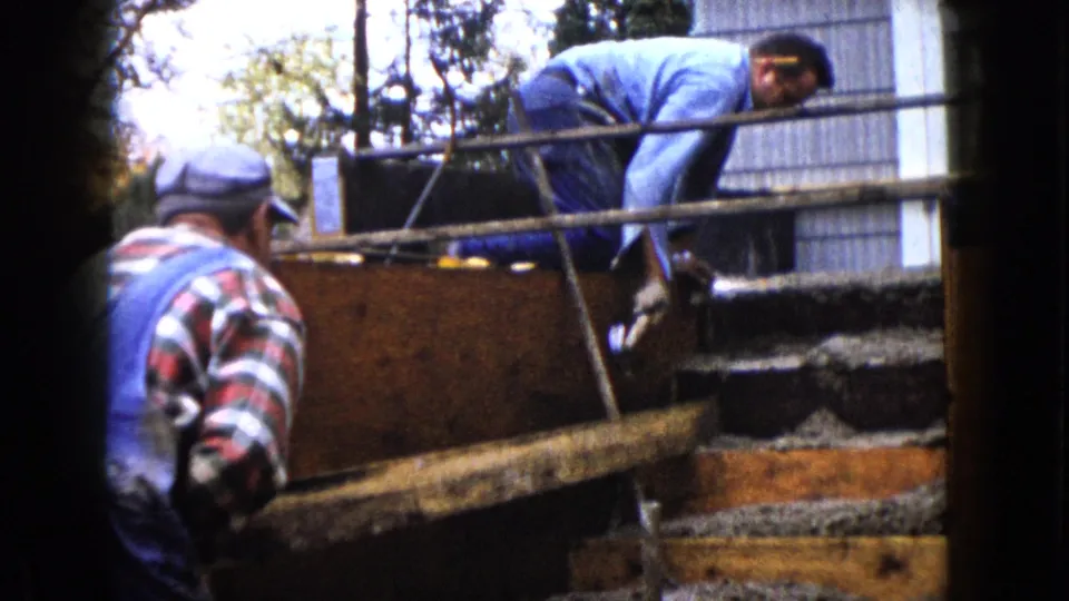 The frame shows a plaid-shirted man close to wooden step forms while a second worker leans over the wet concrete in the background.