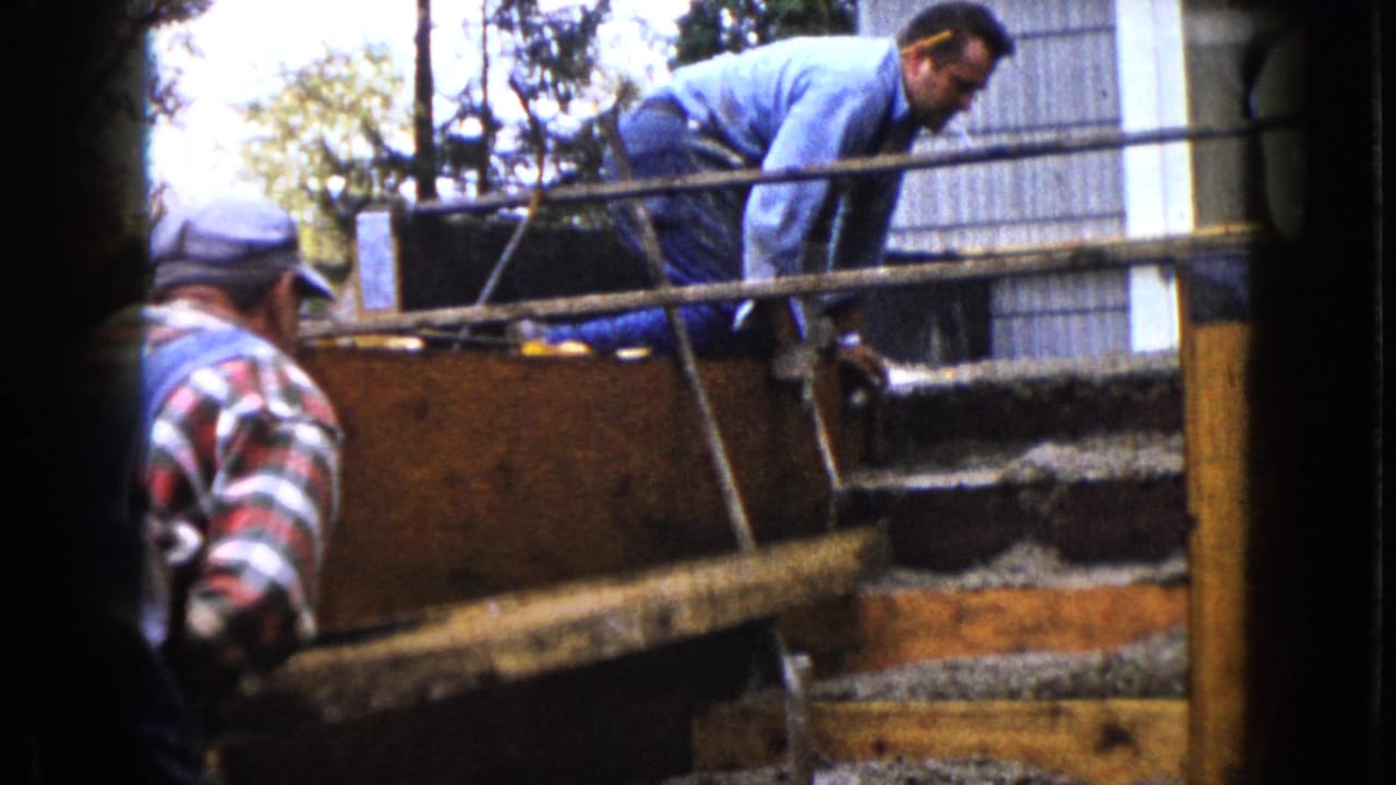 Two men work beside wooden concrete forms at an outdoor construction site.