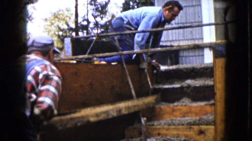 In 1960 Wauconda, Illinois footage, one man leans over a wooden form while another in plaid and overalls stands nearby.