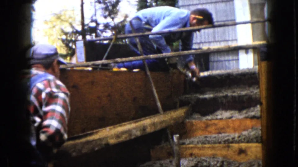 A man in a blue shirt works over wet concrete in wooden forms as another adult in plaid and overalls stands at the left edge.