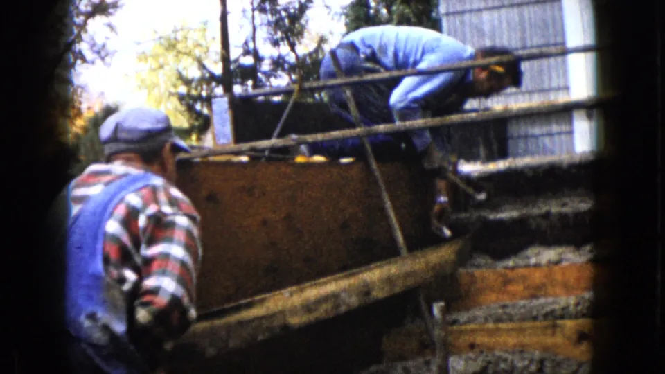 The handheld 8mm frame shows two adults at a partially formed concrete section, with gravel, boards, and wet concrete visible.