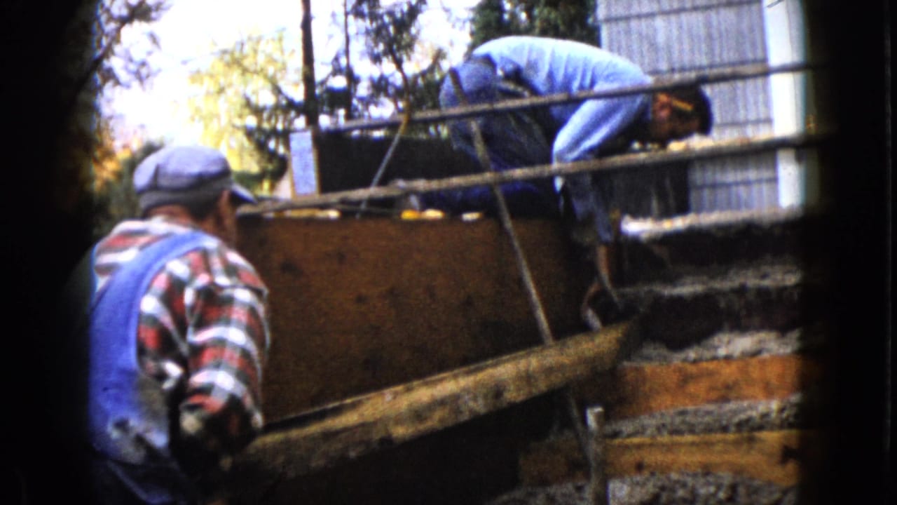 In Wauconda, Illinois, 1960 home-movie footage, two adult men stand near a wooden form during outdoor construction work.
