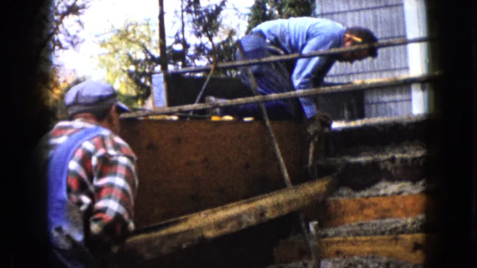 The home-movie frame shows two adult men at a Wauconda construction project, with rough materials and trees behind them.