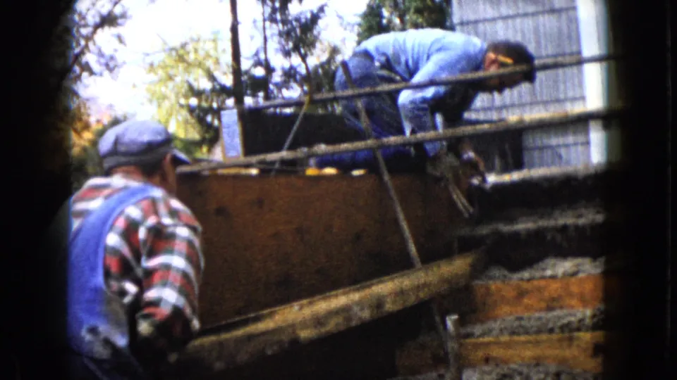A 1960 Wauconda home-movie frame captures one man bent over a wooden construction form as another stands nearby in overalls.