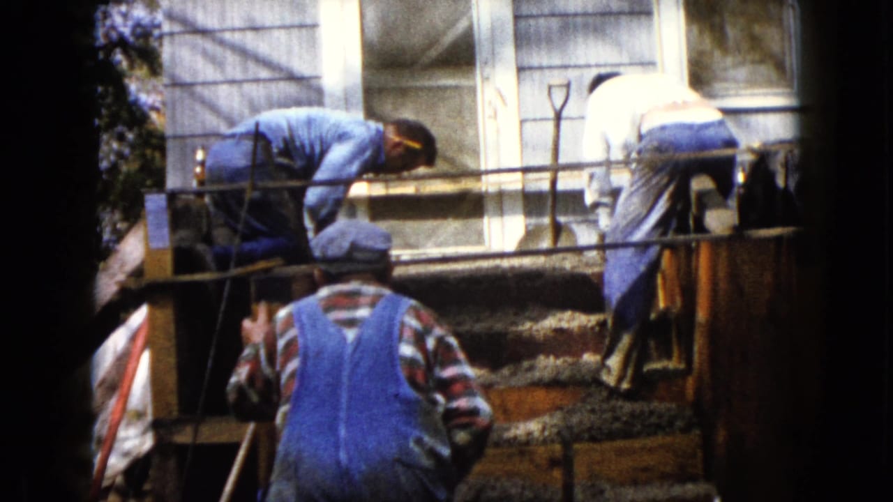 Two men bend over the concrete forms as a third man in blue overalls stands below in this 1960 Wauconda home-movie frame.