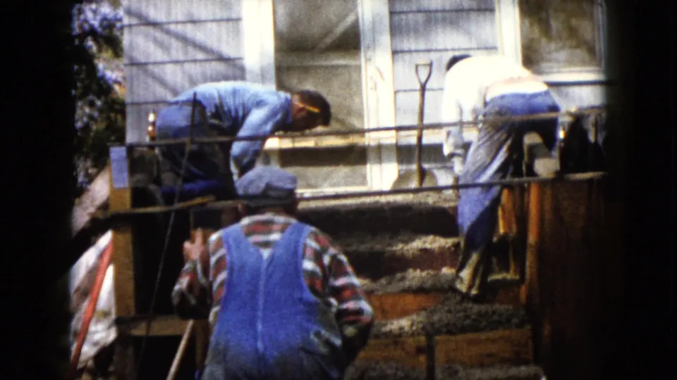 Two men bend over the concrete forms as a third man in blue overalls stands below in this 1960 Wauconda home-movie frame.