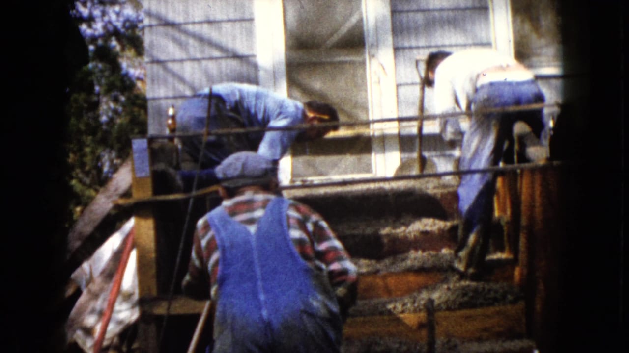 Workers in caps and overalls continue smoothing concrete steps outside a Wauconda house in 1960 home-movie footage.