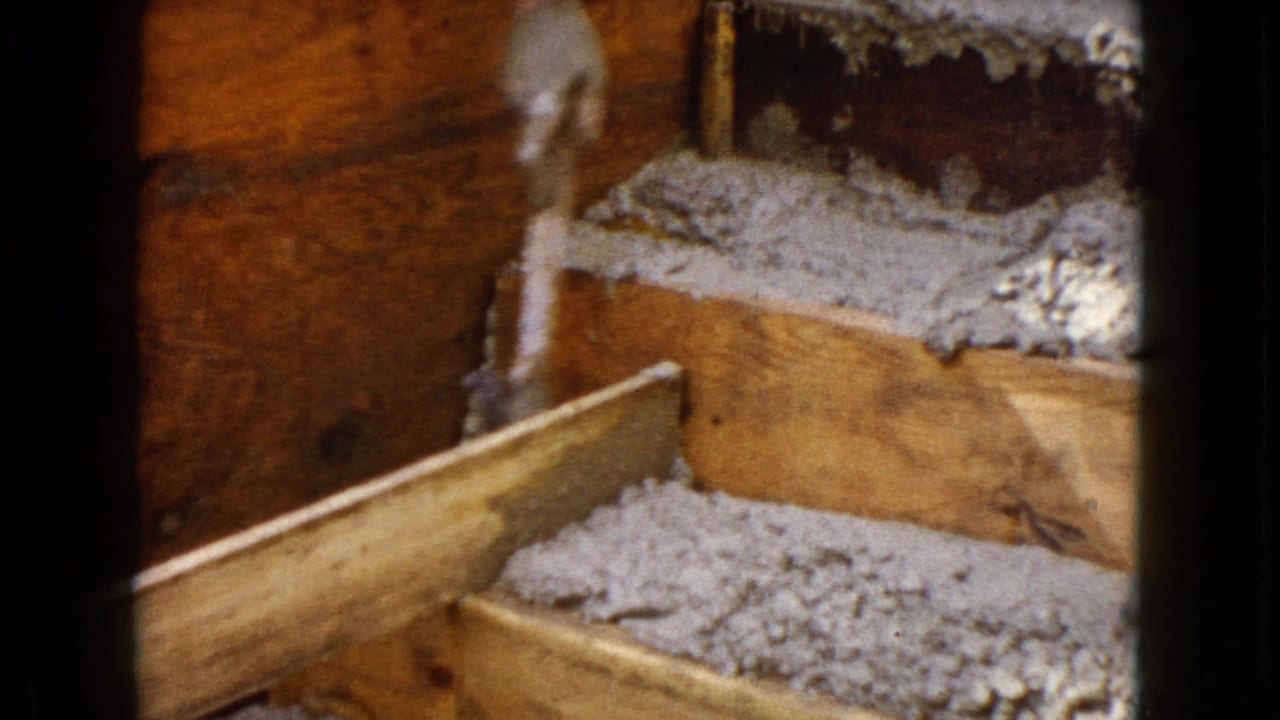 Gloved hand raises a hammer near a wood plank and gray rough material.