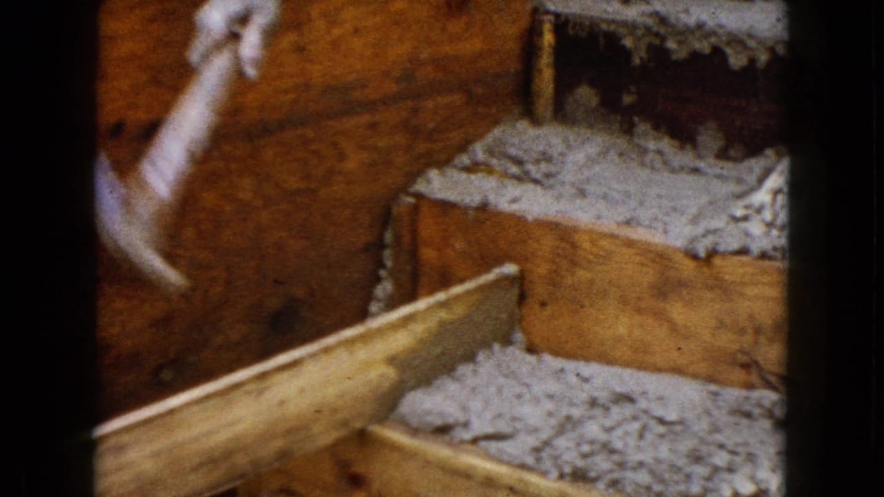 Raised gloved hand and hammer above wood framing with gray material behind.