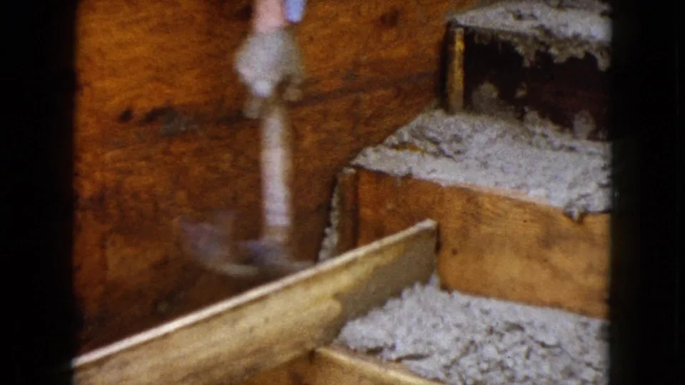 The gloved hand blurs with hammering motion beside a wooden plank and gray aggregate or insulation in 1960 Illinois home-movie footage.