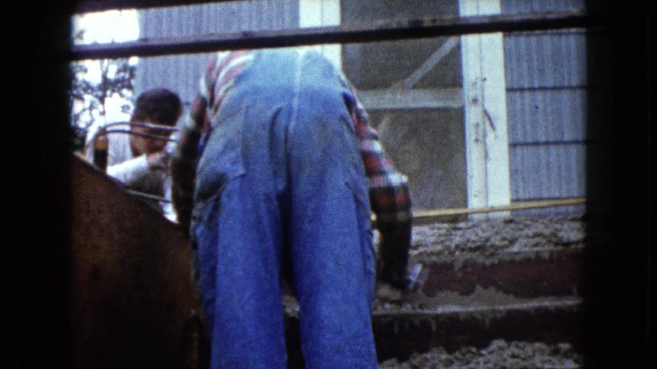 Worker in blue overalls bends over wet cement on exterior stair forms while two men stand nearby.