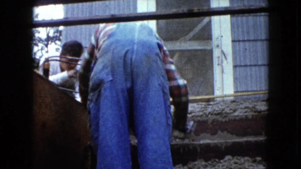 1960 Wauconda, Illinois 8mm frame showing a worker in blue overalls handling wet cement on wooden stair forms outside a house.